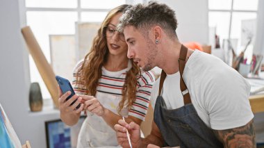 Bir adam ve bir kadın, resim ve malzemelerle dolu bir sanat stüdyosunda akıllı bir telefon kullanıyorlar..