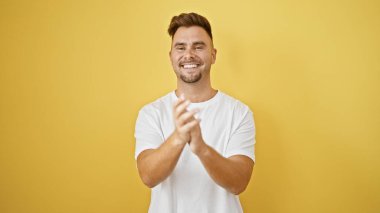 Sakallı, neşeli, genç bir İspanyol adam izole edilmiş sarı bir duvara el çırpıyor.