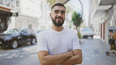 Güneşli bir caddede kolları çapraz duran yakışıklı sakallı adam.