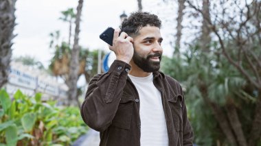 Sakallı yakışıklı genç adam yemyeşil bir parkta akıllı telefon kullanıyor..