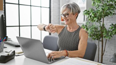 Gülümseyen olgun kadın modern ofiste akıllı saatleri kontrol eder.