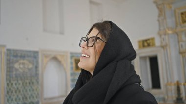 Süslü döşeli tarihi bir Türk sarayında gözlüklü ve başörtüsü takan gülümseyen bir kadın.