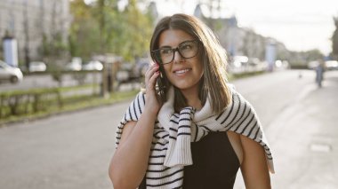 Genç bir İspanyol kadın, güneşli bir Avrupa caddesinde telefonla konuşuyor..