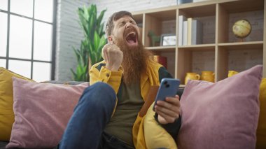 Sakallı adam evde akıllı telefon tutarken heyecanla kutlama yapıyor.