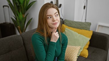 Düşünceli genç bir kadın, rahat bir oturma odasındaki kanepede oturuyor, yakınlarda bir bavulla düşünüyor, seyahat etmeyi öneriyor..