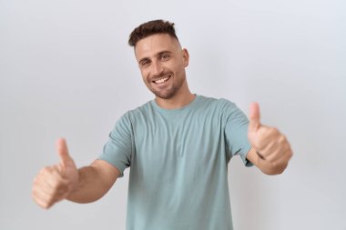 Hispanic man with beard standing over white background approving doing positive gesture with hand, thumbs up smiling and happy for success. winner gesture. 