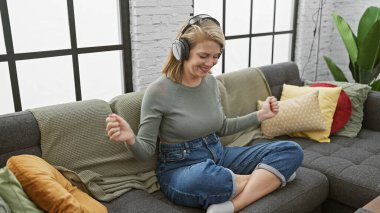 Neşeli genç bir kadın, rahat bir oturma odasındaki kanepede uzanırken kulaklıkla müzik dinlemekten hoşlanır..