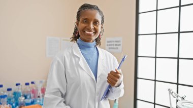 Kendine güvenen Afro-Amerikan kadın bilim adamı. Bir laboratuarda elinde not panosu ve kalemle dikiliyor..
