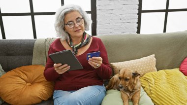 Gözlüklü, tablet kullanan ve kredi kartı tutan olgun bir kadın. Yanında da rahat bir oturma odasında köpeği var..