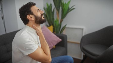 Beyaz tişörtlü sakallı bir adam modern bir oturma odasında boyun ağrısı çekiyor.