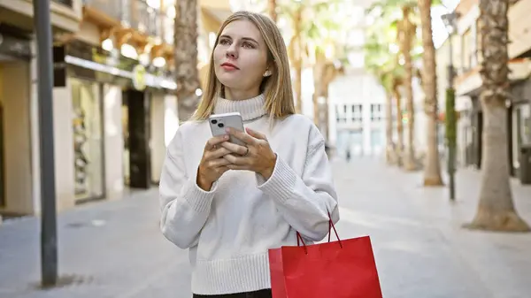一个金发碧眼的女人拿着智能手机和购物袋在城市的街道上 — 图库照片