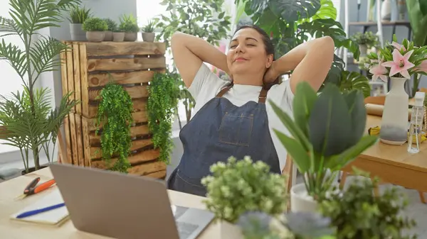 リラックスした中年のヒスパニック女性は 植物と彼女のラップトップに囲まれた緑豊かな屋内花屋で静けさの瞬間を楽しんでいます — ストック写真