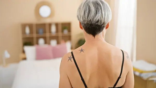Mature woman with grey hair and tattoos in a cozy bedroom setting, embodying tranquility and self-care.
