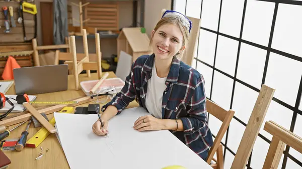 Attractive blonde woman crafts in a sunny workshop, smiling while sketching plans.