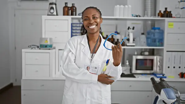 stock image African american woman scientist smiling in a laboratory, holding a pen, with microscope and equipment in the background.