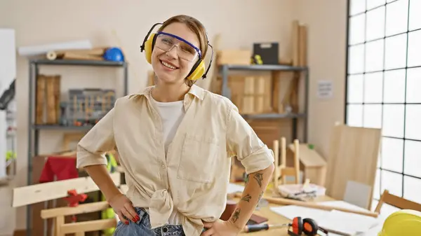Uśmiechnięta Kobieta Okularami Bezpieczeństwa Słuchawkami Stojąca Pewnie Słonecznym Warsztacie Stolarskim — Zdjęcie stockowe