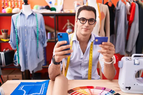 Young hispanic man tailor using smartphone and credit card at atelier