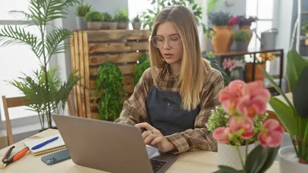 一位年轻的高加索女人在花店里的笔记本电脑上工作 花店四周都是植物和花朵 — 图库照片