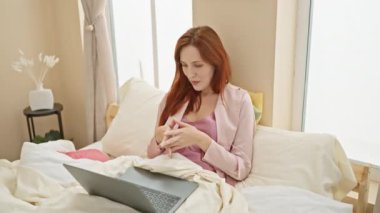 Kızıl saçlı kadın gülümsüyor, yatakta dizüstü bilgisayar kullanıyor, rahat bir yatak odasında, doğal ışık altında..