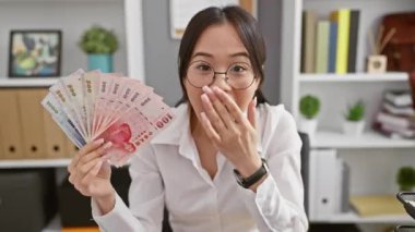 İnançsız genç Çinli bir kadın, ağzı açık, şaşırmış ve korkmuş vaziyette ofisinde Yuan banknotları tutuyor, yüzünde şaşırtıcı bir ifade var.