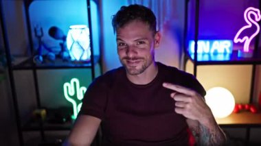 Heyecanlı genç bir adam, sakallı iyimser bir oyuncu, video oyunundaki başarısına gururla işaret ediyor, karanlık bir oyun odasında, kendine güvenen gülümsemesi umutlu bir neşe sunuyor..