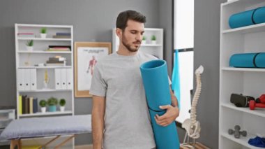 Neşeli, İspanyol asıllı genç adam, yoga minderini kendine güvenle tutuyor, başarısını işaret ederek ifade ediyor, rehabilitasyon kliniğindeki mükemmel gelişmelere gülümsüyor.