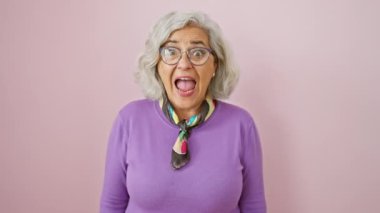 Neşeli orta yaşlı, gri saçlı, gözlüklü, kazanan jestler yapan bir kadın. Heyecanlı kadın zaferin tadını çıkarıyor, pembe izole arka planda başarı çığlıkları atıyor..