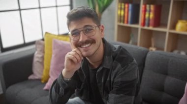 Neşeli, genç İspanyol gözlüklü, dişlek bir gülümseme takan ve evde koltukta güven içinde otururken pozitiflik saçan bir adam..