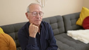 Evde beyaz saçlı endişeli bir adam, oturma odasındaki kanepede oturuyor, ciddi bir yüz ifadesi sergiliyor, şüphe dolu, üzgün ve üzgün, düşüncelerine dalmış.