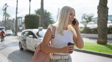 Kendine güvenen genç sarışın kadın mutluluk saçıyor güneşli bir şehir caddesinde sıcak bir kahvenin tadını çıkarırken açık hava yaşam tarzı portresindeki akıllı telefonuna mutlu bir şekilde gülümsüyor.