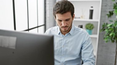 Odaklanmış sakallı İspanyol adam modern ofis ortamında bilgisayar başında çalışıyor, profesyonel ve kentsel yaşam tarzını yansıtıyor..