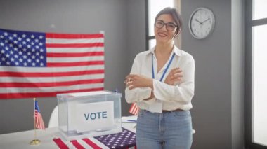 Orta yaşlı, çapraz kollu, kendine güvenen bir İspanyol kadın kapalı bir Birleşik Devletler oy verme merkezinde Amerikan bayrağıyla gülümsüyor..