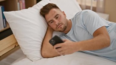 Endişeli genç adam evde akıllı telefonuyla yatakta uzanıyor. Endişelerini ve düşüncelerini dile getiriyor..