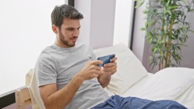 Parlak bir yatak odasında rahatça otururken akıllı telefon kullanan gülümseyen genç adam..