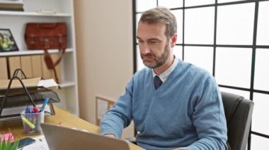 Yakışıklı, orta yaşlı, sakallı, süveterli bir adam. Modern ofis ortamında dizüstü bilgisayarı inceliyor..