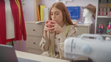 Dikiş malzemeleri ve renkli kumaşlarla dolu bir terzide kupa tutan düşünceli genç bir kadın..