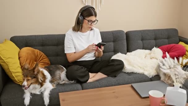 一位心满意足的女人 带着耳机 一边在智能手机上听音乐 一边和睡梦中的狗在沙发上悠闲自在 — 图库视频影像