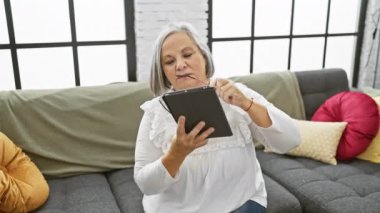 Gri saçlı bir kadın evinin oturma odasında bir tablet okuyor, yaşlıların teknolojik kullanımına örnek olarak..