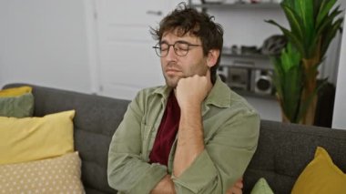 Şık bir oturma odasında gri bir koltukta oturan, gözlüklü ve sakallı düşünceli bir İspanyol.