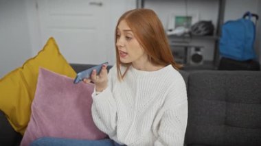 Genç kızıl saçlı bir kadın, rahat oturma odasında renkli yastıklarla birlikte bir sesli mesaj gönderiyor..