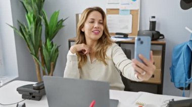 Neşeli bir kadın ofisinde selfie çekiyor, modern bir çalışma alanında arkadaş canlısı, teknoloji meraklısı bir profesyoneli canlandırıyor..
