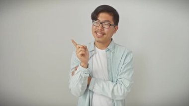 Gözlüklü, güler yüzlü genç Çinli bir adam, güvenli bir şekilde duruyor ve el değmemiş beyaz, izole bir arka planda, dostça bir gülümsemeyle kenarı işaret ediyor.