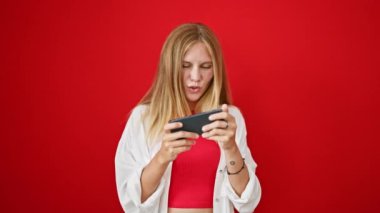 Şaşırmış, sarı saçlı, akıllı telefonunu kırmızı arka planda kullanan genç bir kadın.. 