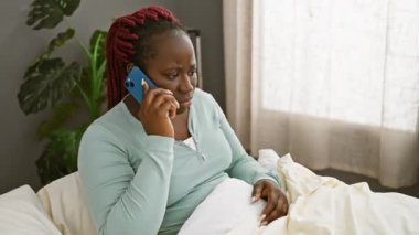 Endişeli bir Afrikalı kadın. Saçları örgülü. Bir klinik odasında, yatakta otururken akıllı bir telefonla konuşuyor..