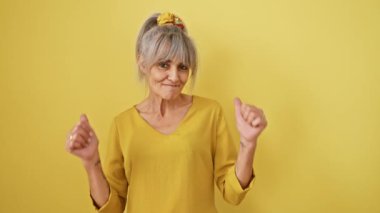 Beyaz saçlı, at kuyruklu neşeli orta yaşlı bir kadın, sarı bir zemine karşı izole bir şekilde duruyor, baş parmak işareti yapıyor. Mutlu bir şekilde gülümsüyor, bir kazananın başarısını yayıyor..