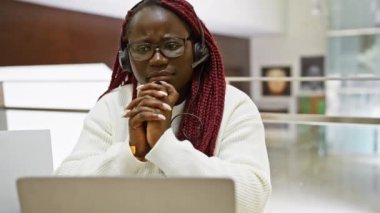 Laptop kullanan ve modern ofis ortamında kulaklık takan düşünceli Afrikalı bir kadın..