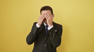Şokta, takım elbiseli genç bir İspanyol adam yüzünü parmaklarından süzerek ürkek bir ifadeyi gözler önüne seriyor, gözlerini gergin bir şekilde gizliyor, güneşli sarı izole edilmiş arka planla çelişiyor..