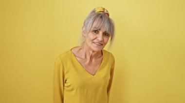 Gri saçlı, neşeli orta yaşlı kadın, komik ve çılgın bir jestle dilini dışarı çıkarıyor. Sarı arka planda izole edilmiş mutlu bir yüz ifadesi sergiliyor.