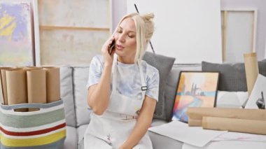 Genç, çekici bir sarışın kadın sanatçının büyüleyici sahnesi, kıkırdayan bir akıllı telefon sohbetine dalmış, rahat bir stüdyo kanepesinde oturuyor sınıf tuvali ve boya fırçaları arasında.