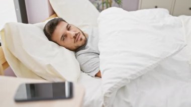 Yatakta yakışıklı genç adam modern bir yatak odası ortamında akıllı telefona uzanıyor.